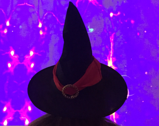 In primo piano un cappello nero da strega, con un nastro rosso alla base e un fermaglio tondo e dorato. Lo sfondo è viola.